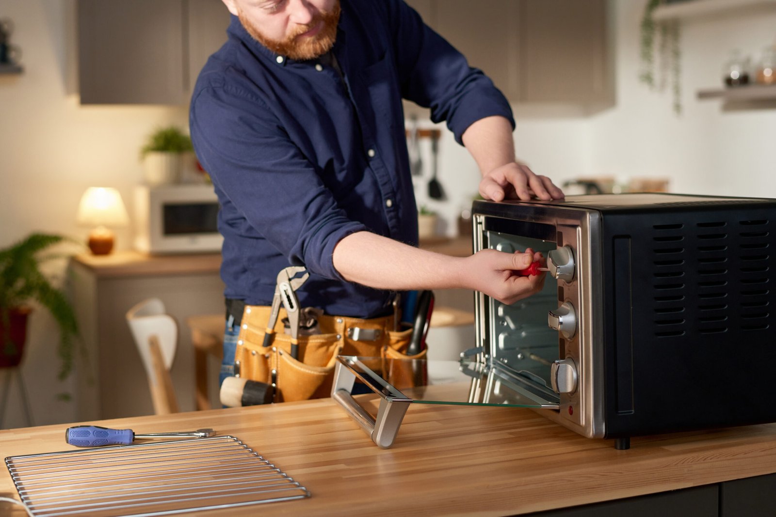 Mature bearded repairman using tools to fix oven at table while standing in domestic kitchen
