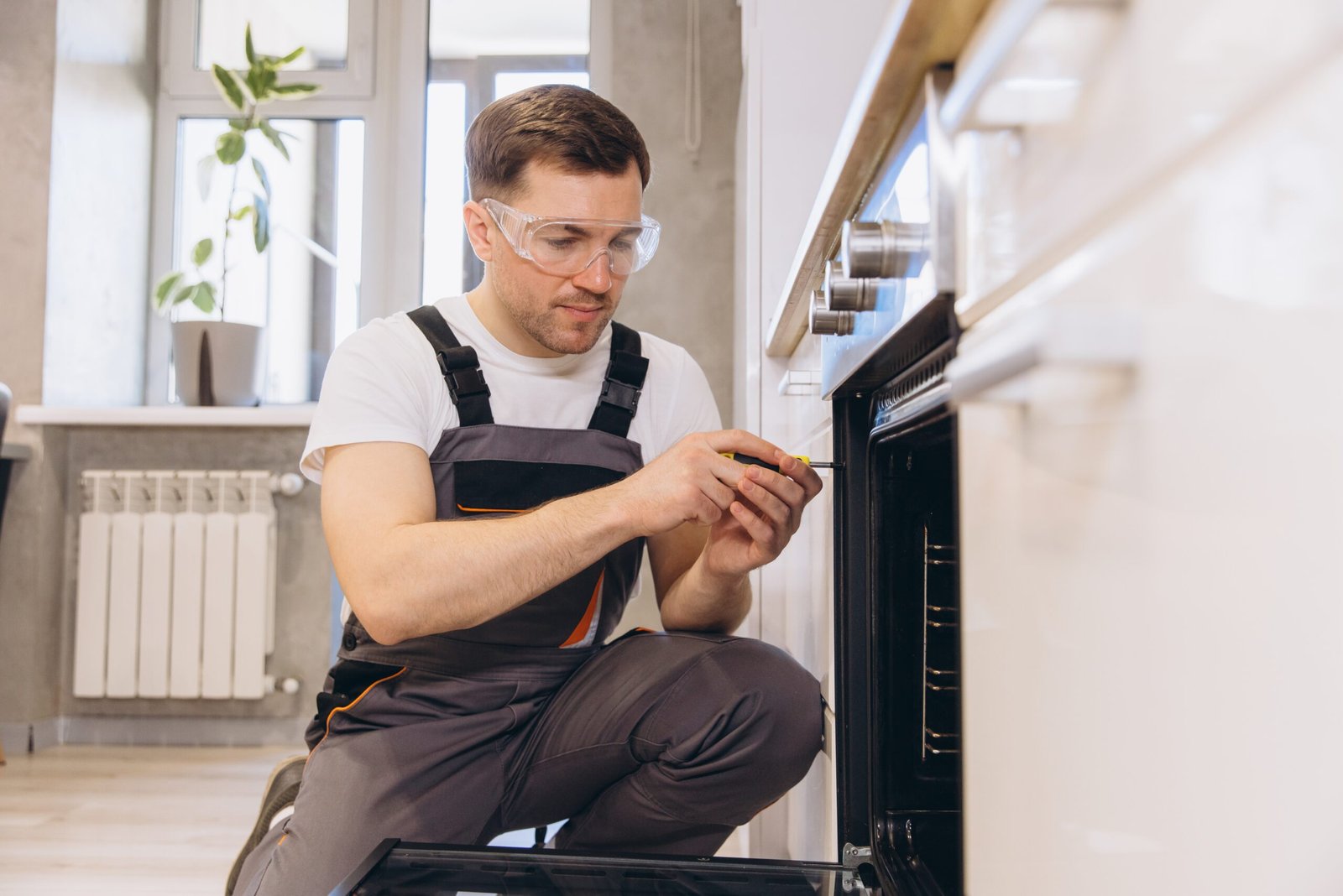Handyman wearing safety glasses and work overalls using a screwdriver to repair a domestic oven in a modern kitchen